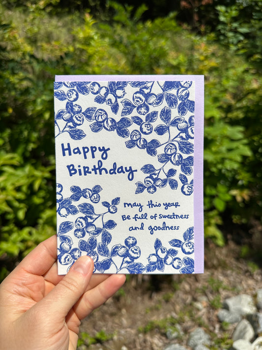 Letterpress greeting card featuring hand-drawn blueberries printed in a vibrant blue ink. "Happy Birthday! May this year be full of sweetness and goodness" is written on the card in a whimsical hand-drawn text, in the same blue ink. The card is white, blank inside, and is paired with a soft lilac envelope. Card is held outside on a sunny summer day.