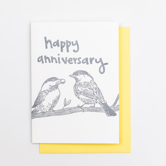 Letterpress greeting card featuring hand-drawn chickadees, printed in a shimmery silver ink. Whimsical hand-drawn text saying "Happy Anniversary" is shown on the top of the card, in the same silver ink. The card is white, blank inside, and is paired with a light yellow envelope.