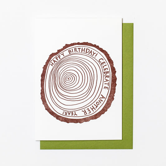 Letterpress greeting card featuring a hand-drawn tree ring printed in an earthy brown ink. Drawn inside one of the outer rings, "Happy Birthday! Celebrate another year" is written in a whimsical hand-drawn text, in the same earthy brown ink. The card is white, blank inside, and is paired with a green envelope.