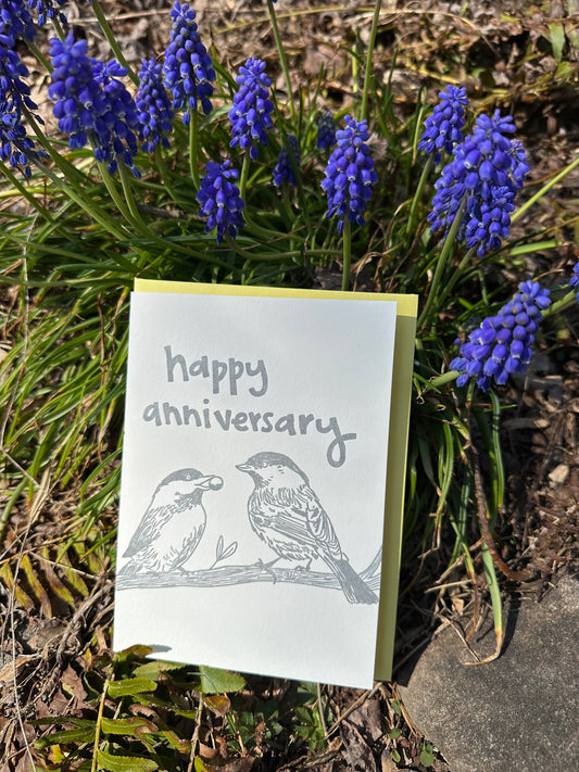 Letterpress greeting card featuring hand-drawn chickadees, printed in a shimmery silver ink. Whimsical hand-drawn text saying "Happy Anniversary" is shown on the top of the card, in the same silver ink. The card is white, blank inside, and is paired with a light yellow envelope. Card is shown outside on a sunny day in front of grape hyacinth flowers.