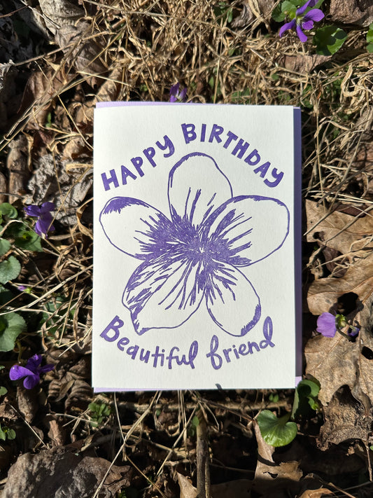 Letterpress greeting card featuring a hand-drawn Violet flower printed in a vibrant purple ink. "Happy Birthday Beautiful Friend!" is written on the top and bottom of the flower in a whimsical hand-drawn text, in the same purple ink. The card is white, blank inside, and is paired with a lilac purple envelope. Card is shown outside on the ground atop some violets on a sunny winter day.