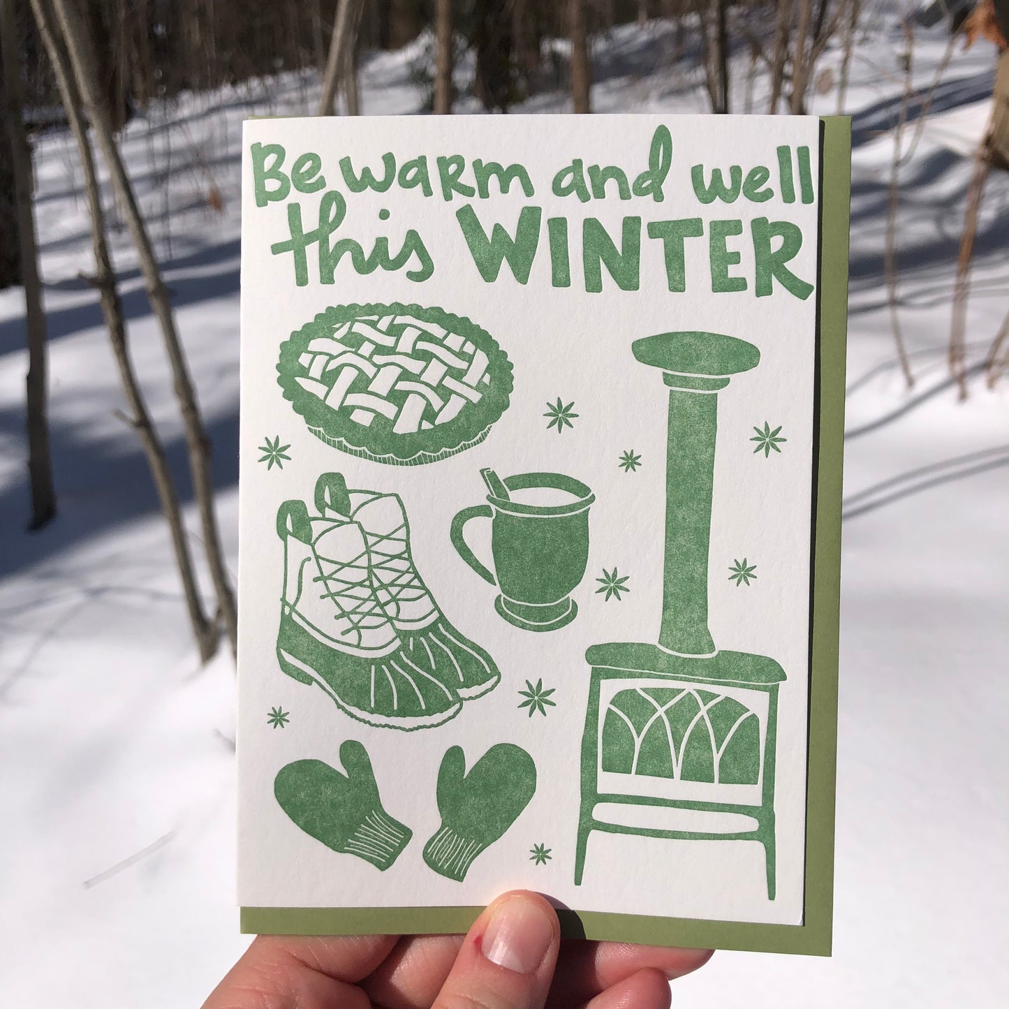 Celebrate Winter with this festive greeting card! This cheerful and cozy card features all the iconic image of the winter season: wood burning stove, star anise, hot cocoa (or cider!), fresh baked pie, mittens, winter boots. Hand-drawn images and typography are letterpress-printed in forest green ink. Green envelope. Card is shown outside in the winter snow.
