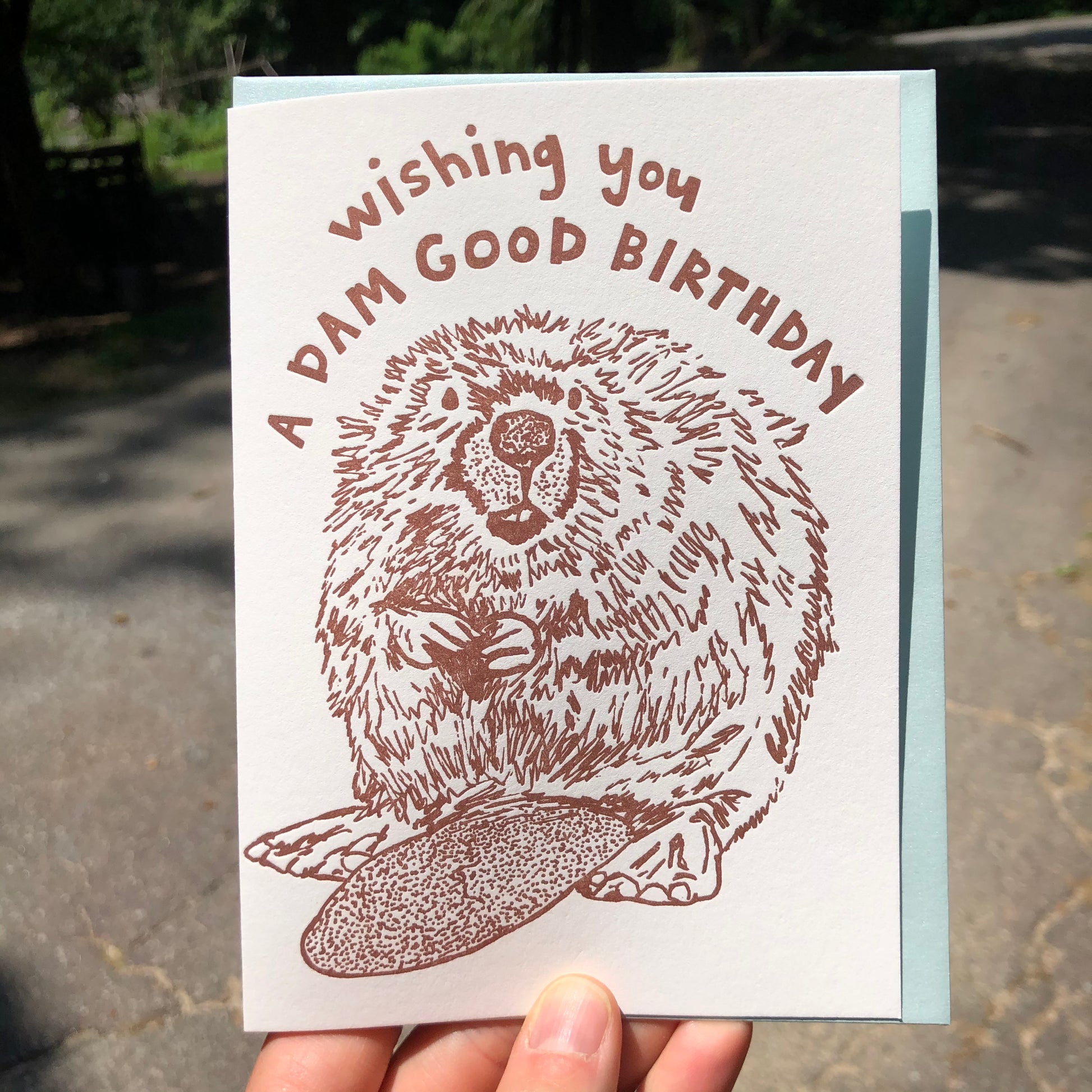 Letterpress greeting card featuring a hand-drawn Beaver printed in an earthy brown ink. "Wishing you a Dam Good Birthday!" is written on the top above the beaver in a whimsical hand-drawn text, in the same brown ink. The card is white, blank inside, and is paired with a light sky blue envelope. Card is being held up outside on a sunny day.