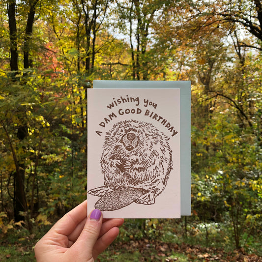 Letterpress greeting card featuring a hand-drawn Beaver printed in an earthy brown ink. "Wishing you a Dam Good Birthday!" is written on the top above the beaver in a whimsical hand-drawn text, in the same brown ink. The card is white, blank inside, and is paired with a light sky blue envelope. Card is being help up in front of an early Autumn deciduous forest.