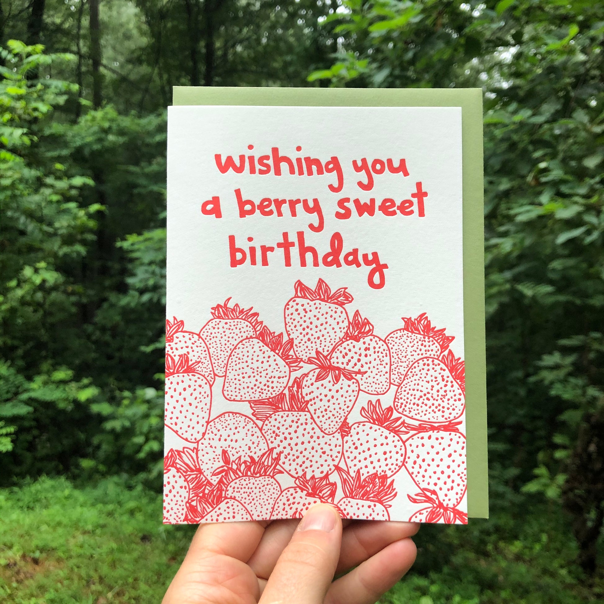 Letterpress greeting card featuring hand-drawn strawberries printed in a vibrant red ink. "Wishing you a Berry Sweet Birthday!" is written at the top of the card in a whimsical hand-drawn text, printed in the same red ink. The card is white, blank inside, and is paired with a deep green envelope. Card is held in front of a lush green summer forest outside.