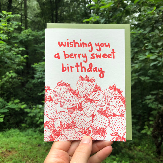 Letterpress greeting card featuring hand-drawn strawberries printed in a vibrant red ink. "Wishing you a Berry Sweet Birthday!" is written at the top of the card in a whimsical hand-drawn text, printed in the same red ink. The card is white, blank inside, and is paired with a deep green envelope. Card is held in front of a lush green summer forest outside.