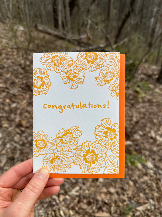 Letterpress greeting card featuring cheerful Coreopsis flowers, printed in a vibrant golden-orange ink. Whimsical hand-drawn text saying "congratulations" is shown in the center of the card, in the same golden-orange ink. The card is white, blank inside, and is paired with a fun orange envelope. Card is shown in a winter forest.