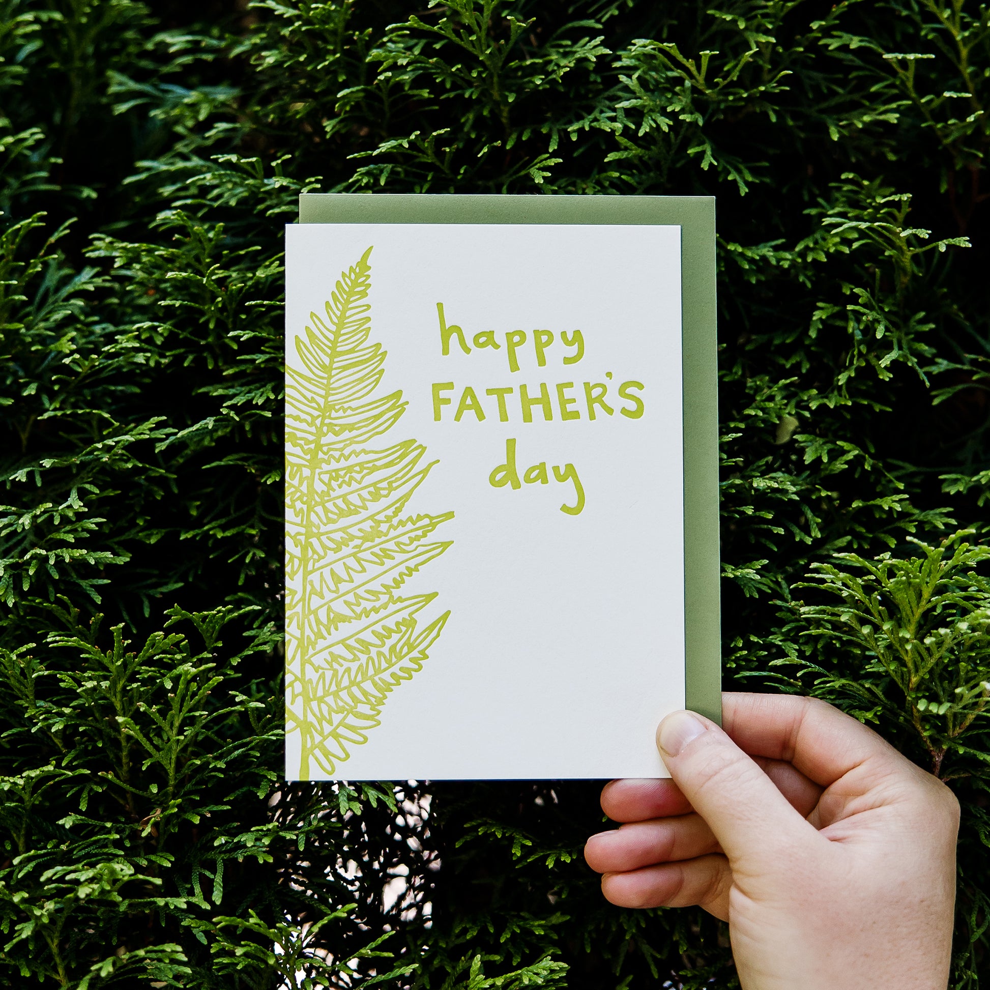 Letterpress greeting card featuring a hand-drawn fern, printed in a vibrant lime green ink. The top right of the card says "Happy Father's Day" in a whimsical hand-drawn text, in the same lime green ink. The card is white, blank inside, and is paired with a green envelope. Card is held in front of a green hedge, zoomed in.