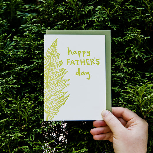 Letterpress greeting card featuring a hand-drawn fern, printed in a vibrant lime green ink. The top right of the card says "Happy Father's Day" in a whimsical hand-drawn text, in the same lime green ink. The card is white, blank inside, and is paired with a green envelope. Card is held in front of a green hedge, zoomed in.