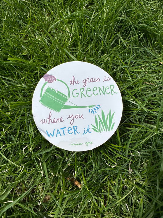 A 4" x 4" vinyl sticker featuring the words: The Grass is Greener Where You Water It" and a hand-drawn plum hand holding a green watering can, pouring blue water droplets on a patch of leafy grass. The text and original artwork hand-drawn by Macon York in her funky folk-art style. The sticker features three colors: a vibrant fern green, a rich plum, and a deep sky blue. Sticker is shown outside on the grass.