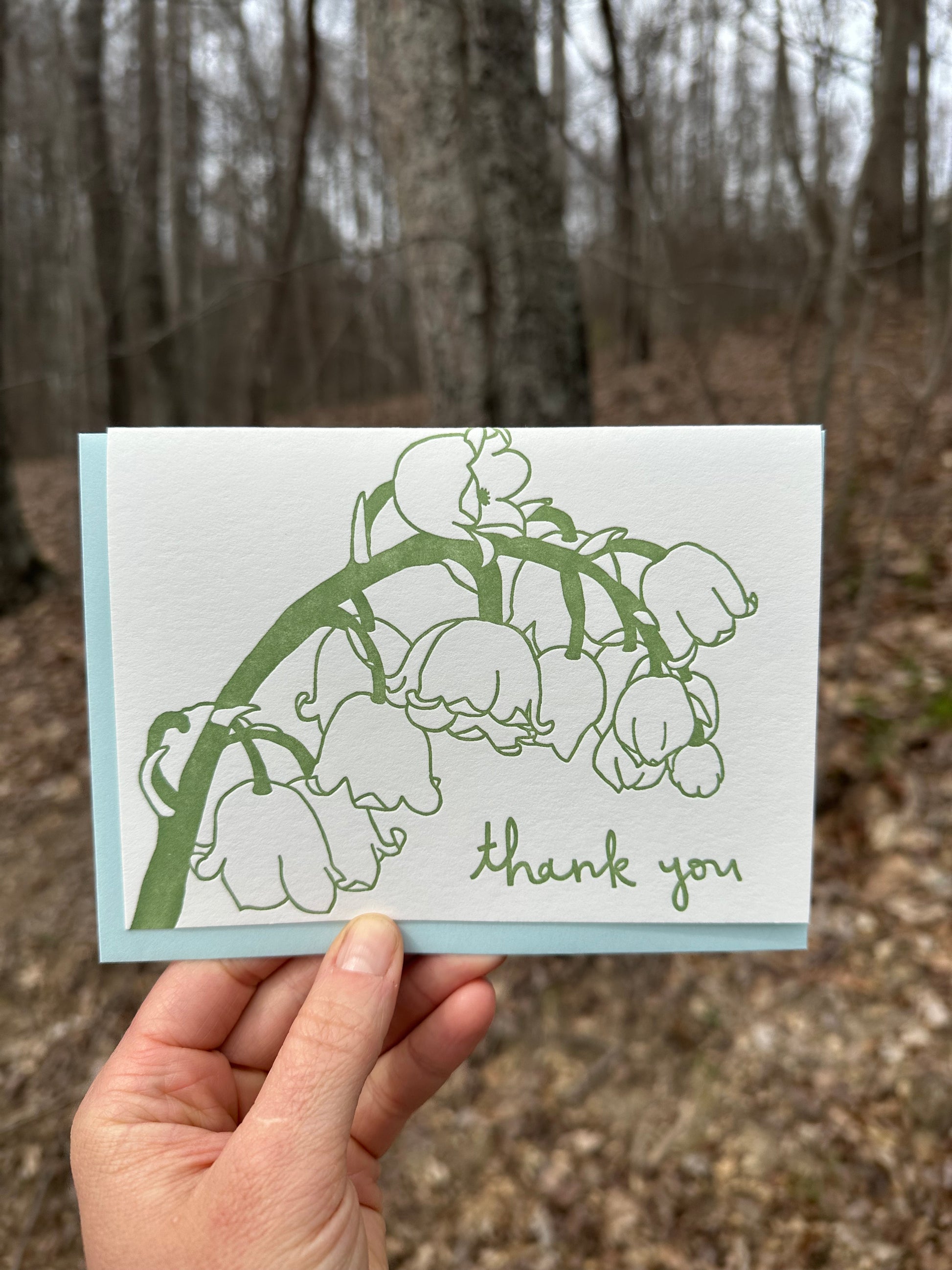 Letterpress greeting card featuring hand-drawn Lily of the Vlley, printed in a rich sage green ink. Whimsical hand-drawn text saying "Thank you" is shown at the bottom right side of the card, in the same green ink. The card is white, blank inside, and is paired with a light sky blue envelope. Card is shown outside in front of a deciduous forest in the winter.