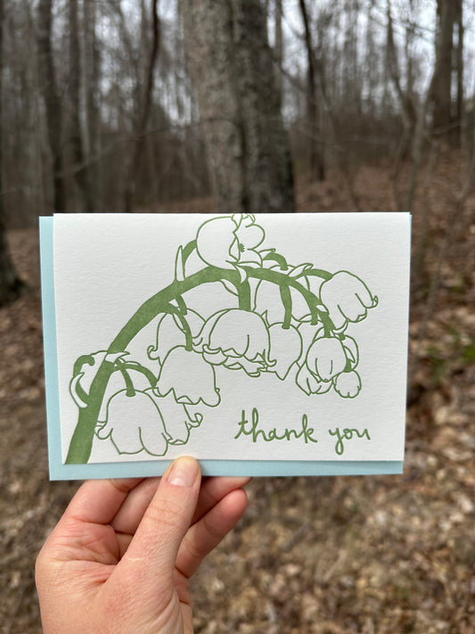 Letterpress greeting card featuring hand-drawn Lily of the Vlley, printed in a rich sage green ink. Whimsical hand-drawn text saying "Thank you" is shown at the bottom right side of the card, in the same green ink. The card is white, blank inside, and is paired with a light sky blue envelope. Card is shown outside in front of a deciduous forest in the winter.