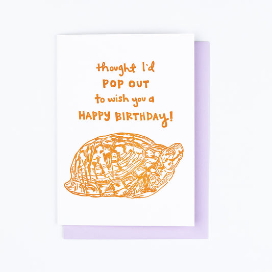 Letterpress greeting card featuring a hand-drawn Appalachian Box Turtle printed in a vibrant dark orange ink. "Thought I'd pop out to wish you a Happy Birthday!" is written on the top of the card in a whimsical hand-drawn text, in the same orange ink. The card is white, blank inside, and is paired with a lilac purple envelope.