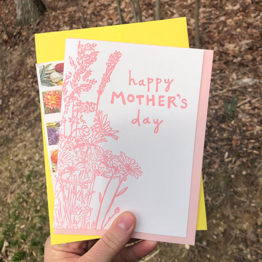 Letterpress greeting card featuring a hand-drawn wildflower bouquet, printed in a vibrant pink ink. The top center of the card says "Happy Mother's Day" in a whimsical hand-drawn text, in the same pink ink. The card is white, blank inside, and is paired with a light pink envelope.