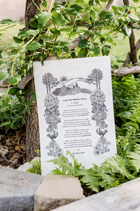Large letterpress art print featuring "The Old North State," an ode to North Carolina and all her natural beauty, written in 1904 by Mrs. Leonora Monteiro. Macon York has hand-written the entire toast in her whimsical style. All the native plants in the toast are hand-drawn to create a border around the text.  The print is shown outside behind some rocks, in front of an arbor with honeysuckle growing up, and wild ferns to the right.