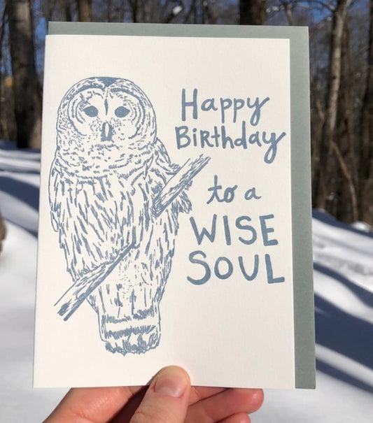 Letterpress greeting card featuring a hand-drawn barred owl printed in a lovely navy ink. The right side of the card says "Happy Birthday to a wise soul!" in a whimsical hand-drawn text, in the same navy ink. The card is white, blank inside, and is paired with a slate grey envelope. Card is shown outside in a snowy forest.