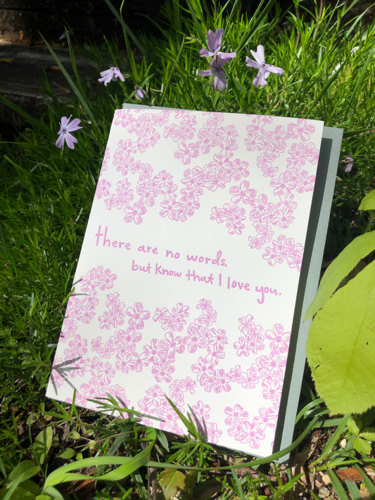 Letterpress greeting card featuring hand-drawn phlox flowers, printed in vibrant lavender ink. Whimsical hand-drawn text saying "There are no words, but know that I love you" is shown in the center of the card, in the same lavender ink. The card is white, blank inside, and is paired with a slate gray envelope. The card is shown outside with light purple phlox on a sunny spring day.