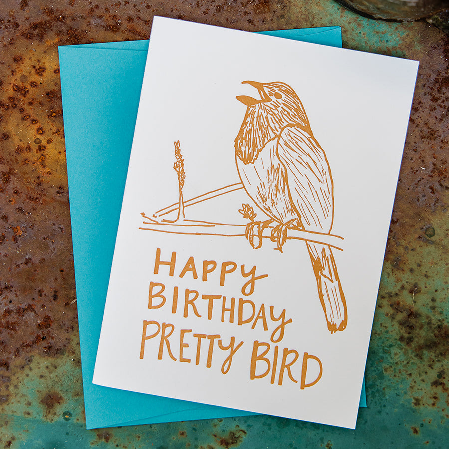 Letterpress greeting card featuring a hand-drawn Eastern towhee printed in a vibrant dark orange ink. The bottom of the card says "Happy Birthday Pretty Bird!" in a whimsical hand-drawn text, in the same dark orange ink. The card is white, blank inside, and is paired with a deep turquoise envelope. Card is on a rusty turquoise outdoor table.