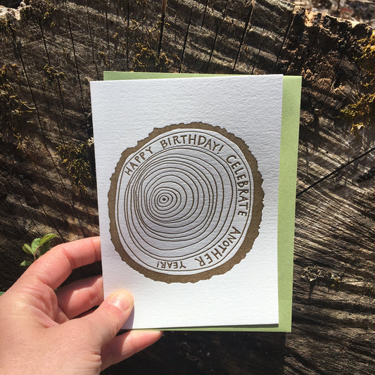 Letterpress greeting card featuring a hand-drawn tree ring printed in an earthy brown ink. Drawn inside one of the outer rings, "Happy Birthday! Celebrate another year" is written in a whimsical hand-drawn text, in the same earthy brown ink. The card is white, blank inside, and is paired with a green envelope. Card shown in front of a cross section of a tree log.