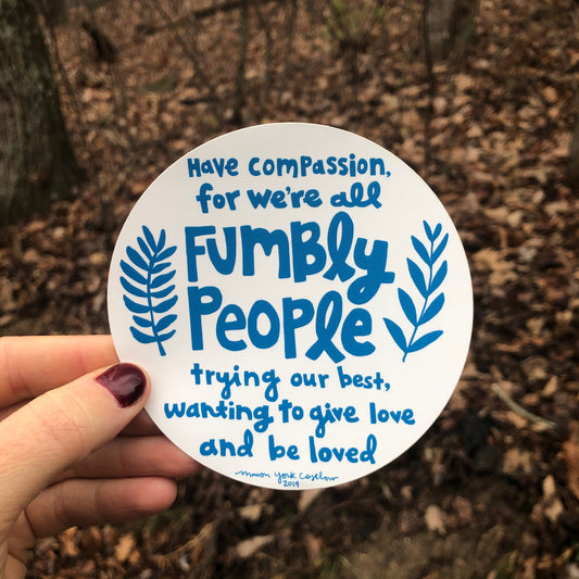This vinyl sticker features original text by Macon York, hand-drawn in a funky folk-art style. 4" x 4" Circle Vinyl Sticker. Text reads "Have compassion, for we're all fumbly people trying our best, wanting to give love and be loved." Sticker is shown outside in front of a winter forest.