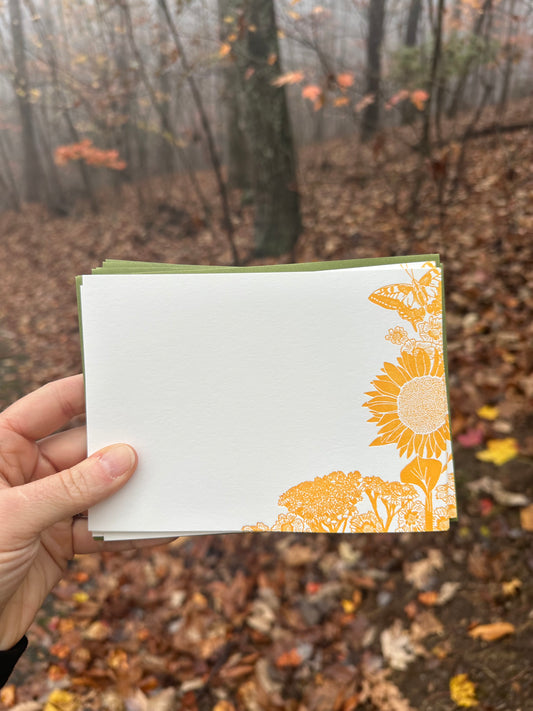 Hand holding a letterpress note card featuring golden-yellow illustrations of a sunflower, wildflowers, and a butterfly. Paired with a soft green envelope. Background shows a misty autumn forest with fallen leaves.