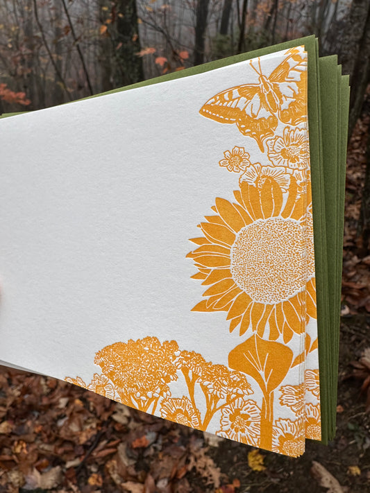 Hand holding a letterpress note card featuring golden-yellow illustrations of a sunflower, wildflowers, and a butterfly. Paired with a soft green envelope. Background shows a misty autumn forest with fallen leaves.