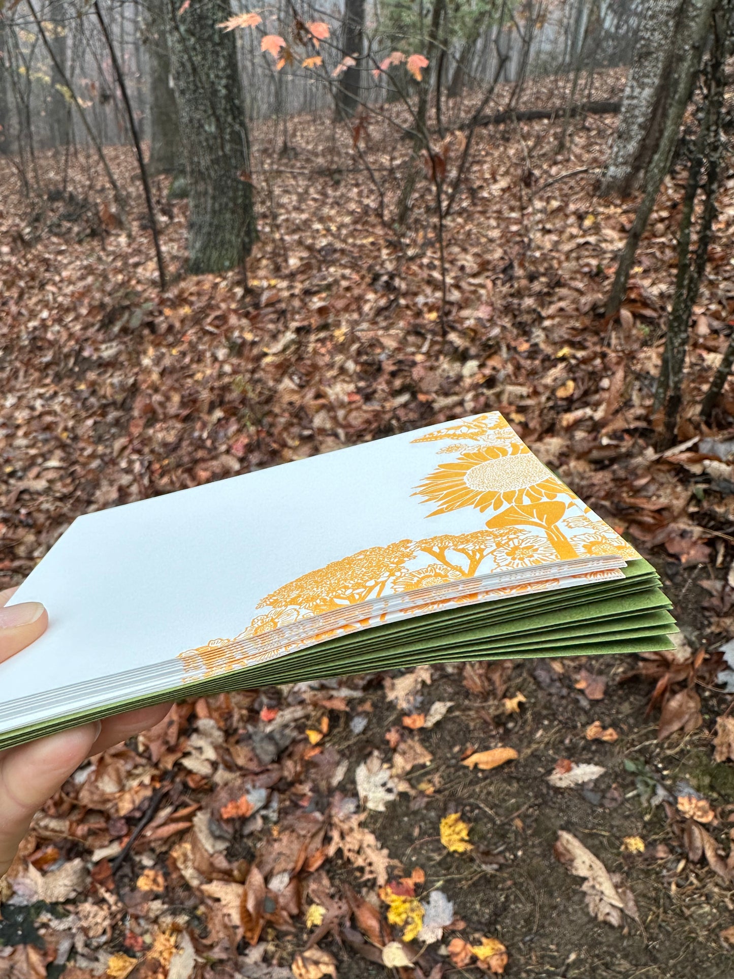 Hand holding a letterpress note card featuring golden-yellow illustrations of a sunflower, wildflowers, and a butterfly. Paired with a soft green envelope. Background shows a misty autumn forest with fallen leaves.