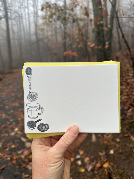 "Hand holding a letterpress note card with hand-drawn black illustrations of a wooden spoon, onion, measuring cup, and vegetables. Paired with a golden-yellow envelope, the background shows a misty autumn forest."