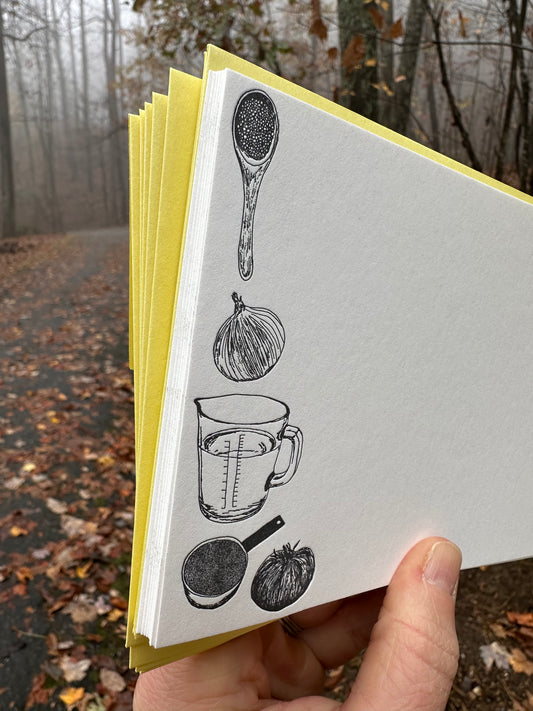 Hand holding a letterpress note card with hand-drawn black illustrations of a wooden spoon, onion, measuring cup, and vegetables. Paired with a golden-yellow envelope, the background shows a misty autumn forest.