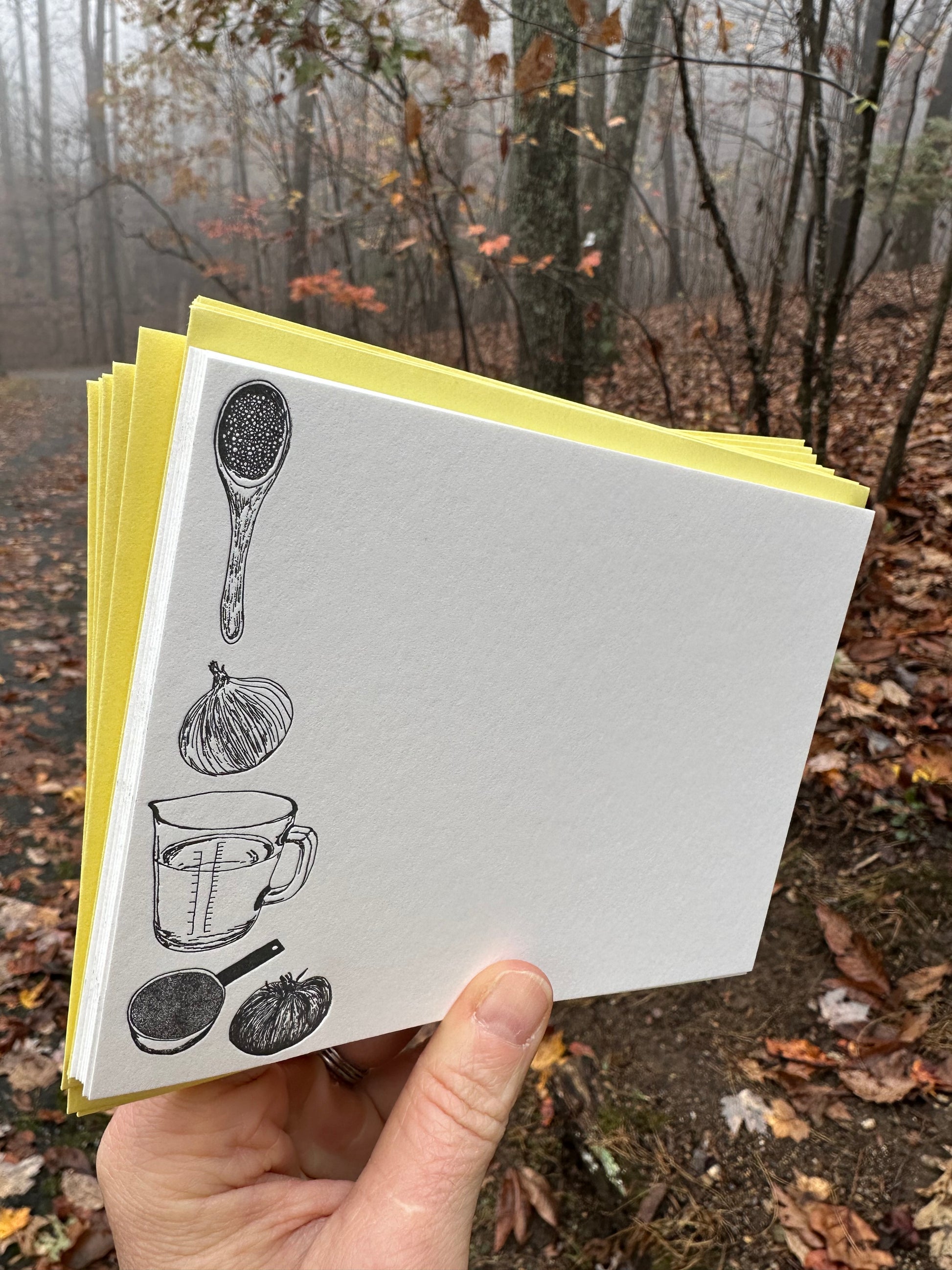 "Hand holding a letterpress note card with hand-drawn black illustrations of a wooden spoon, onion, measuring cup, and vegetables. Paired with a golden-yellow envelope, the background shows a misty autumn forest."