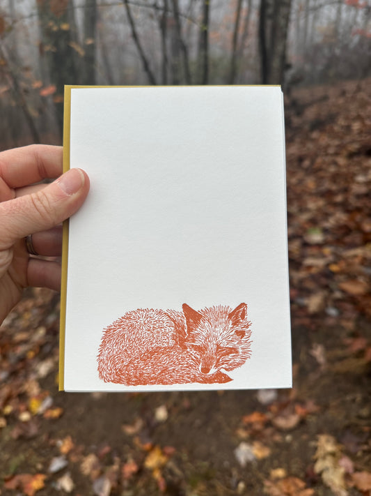 Hand holding a vertically oriented letterpress note card featuring a rust-orange illustration of a curled-up sleeping fox. Paired with a golden-ochre envelope. The background shows a misty autumn forest with fallen leaves.