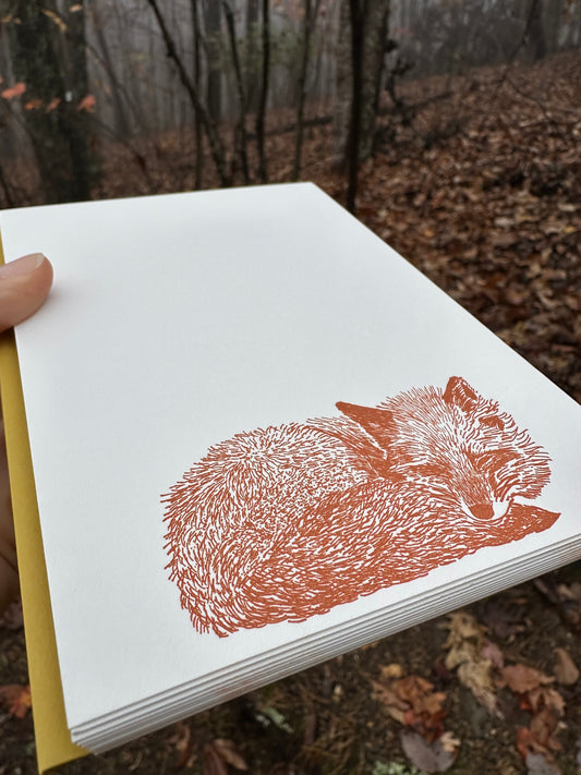 Hand holding a vertically oriented letterpress note card featuring a rust-orange illustration of a curled-up sleeping fox. Paired with a golden-ochre envelope. The background shows a misty autumn forest with fallen leaves.