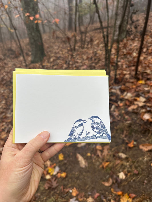 Hand holding a letterpress note card featuring two hand-drawn chickadees perched on a branch, printed in deep navy ink. Paired with a golden-yellow envelope. The background shows a misty forest with fallen leaves.