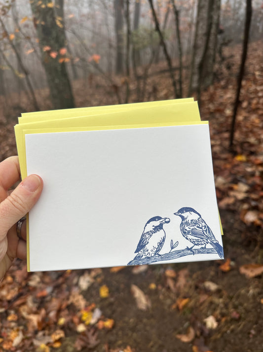 Hand holding a letterpress note card featuring two hand-drawn chickadees perched on a branch, printed in deep navy ink. Paired with a golden-yellow envelope. The background shows a misty forest with fallen leaves.