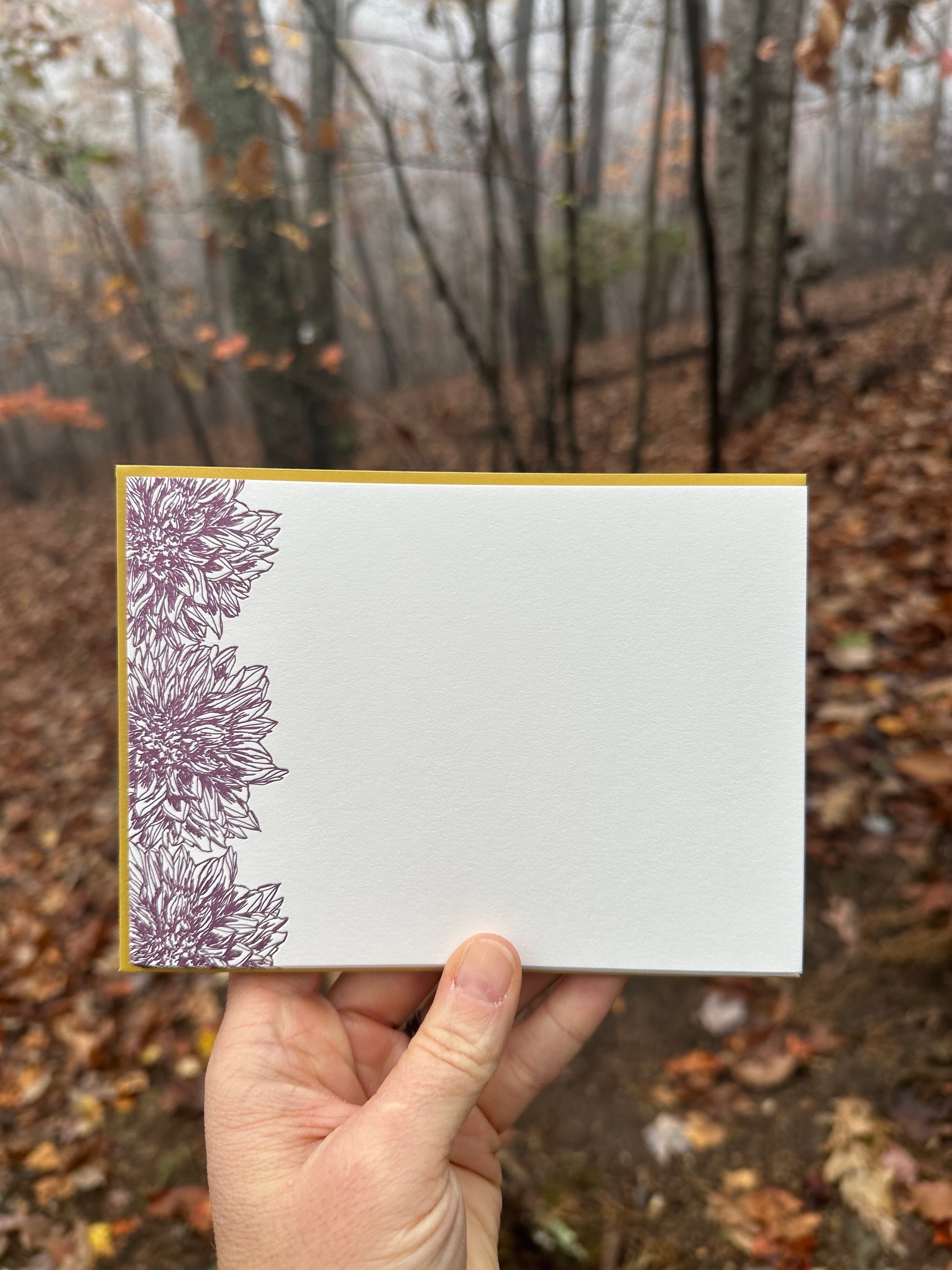 Hand holding a letterpress note card featuring intricate deep purple dahlia illustrations on the left edge, paired with a warm golden-ochre envelope. The background shows a misty autumn forest with fallen leaves and bare trees.