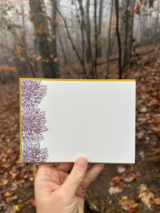 Hand holding a letterpress note card featuring intricate deep purple dahlia illustrations on the left edge, paired with a warm golden-ochre envelope. The background shows a misty autumn forest with fallen leaves and bare trees.