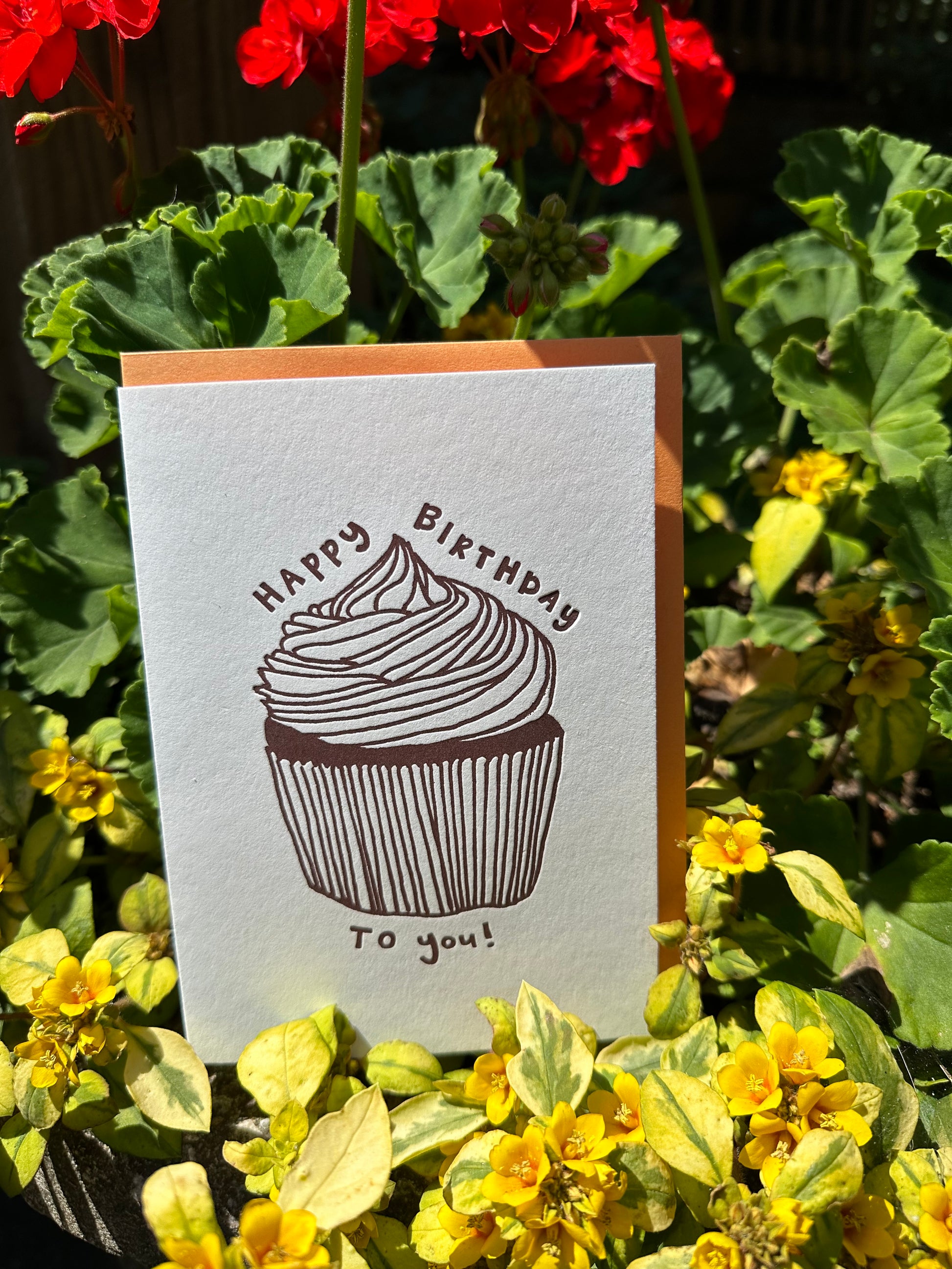 Letterpress greeting card featuring a hand-drawn cupcake printed in a chocolate brown ink. "Happy Birthday to you!" is written around the cupcake in a whimsical hand-drawn text, in the same brown ink. The card is white, blank inside, and is paired with a soft orange envelope. Card is shown with red geraniums and vining plant with yellow flowers, outside on a sunny summer day. 