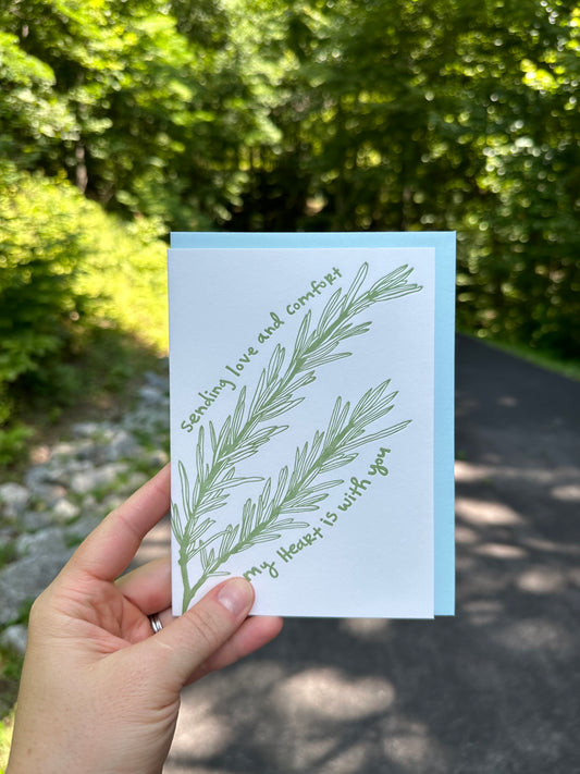 Letterpress greeting card featuring an intricate hand-drawn rosemary herb sprig, printed in a rich sage green ink. Whimsical hand-drawn text saying "Sending love and comfort. My heart is with you." is printed in the same green ink. The card is white, blank inside, and is paired with a light blue envelope. The card is shown outside on a country road, on a sunny summer day. 