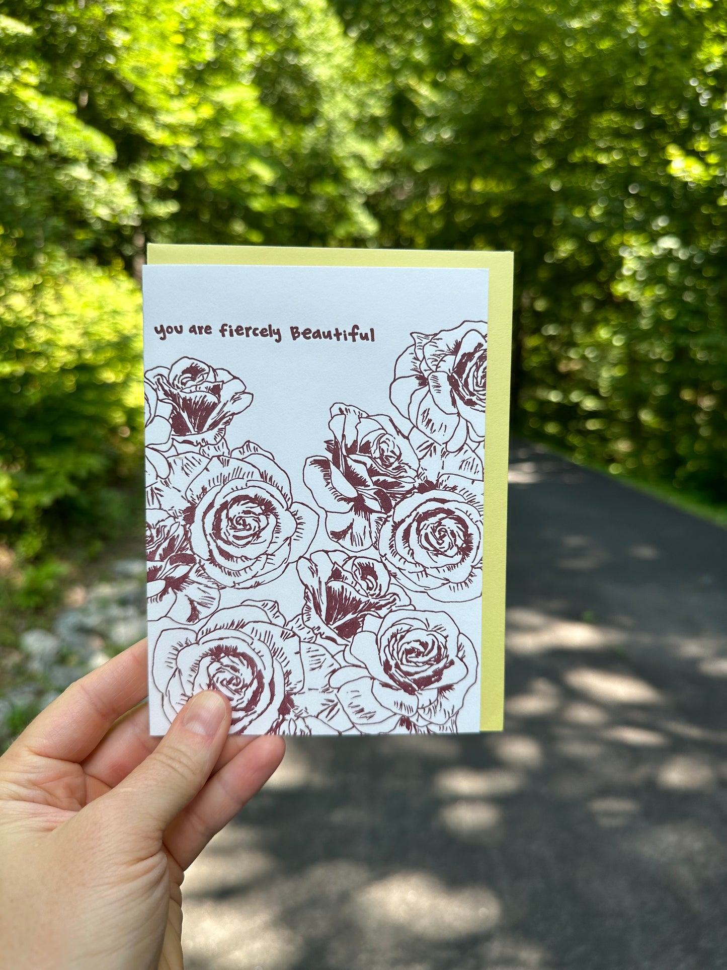 Letterpress greeting card featuring hand-drawn rose flowers printed in a rich, warm brown ink. Whimsical hand-drawn text saying "you are fiercely beautiful" is shown at the top of the card, in the same brown ink. The card is white, blank inside, and is paired with a soft yellow envelope. Card is shown outside in front of a country rode on a sunny summer day. 