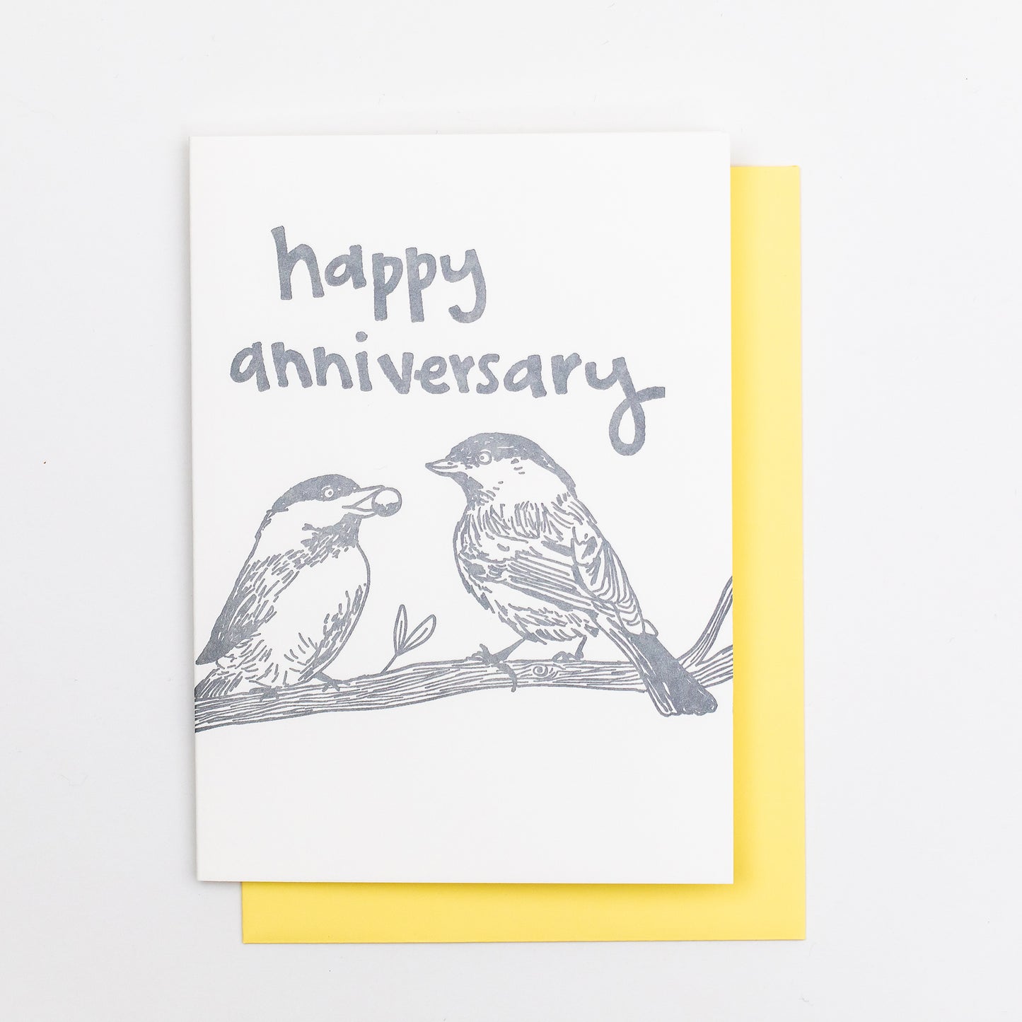 Letterpress greeting card featuring hand-drawn chickadees, printed in a shimmery silver ink. Whimsical hand-drawn text saying "Happy Anniversary" is shown on the top of the card, in the same silver ink. The card is white, blank inside, and is paired with a light yellow envelope.