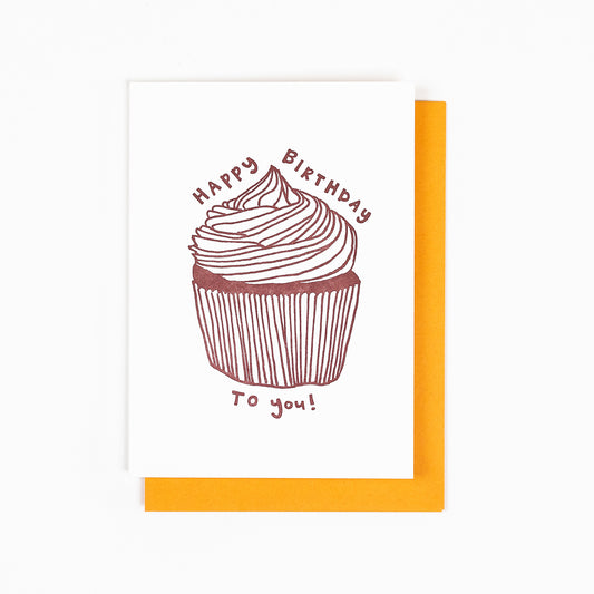 Letterpress greeting card featuring a hand-drawn cupcake printed in a chocolate brown ink. "Happy Birthday to you!" is written around the cupcake in a whimsical hand-drawn text, in the same brown ink. The card is white, blank inside, and is paired with a soft orange envelope.