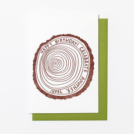 Letterpress greeting card featuring a hand-drawn tree ring printed in an earthy brown ink. Drawn inside one of the outer rings, "Happy Birthday! Celebrate another year" is written in a whimsical hand-drawn text, in the same earthy brown ink. The card is white, blank inside, and is paired with a green envelope.