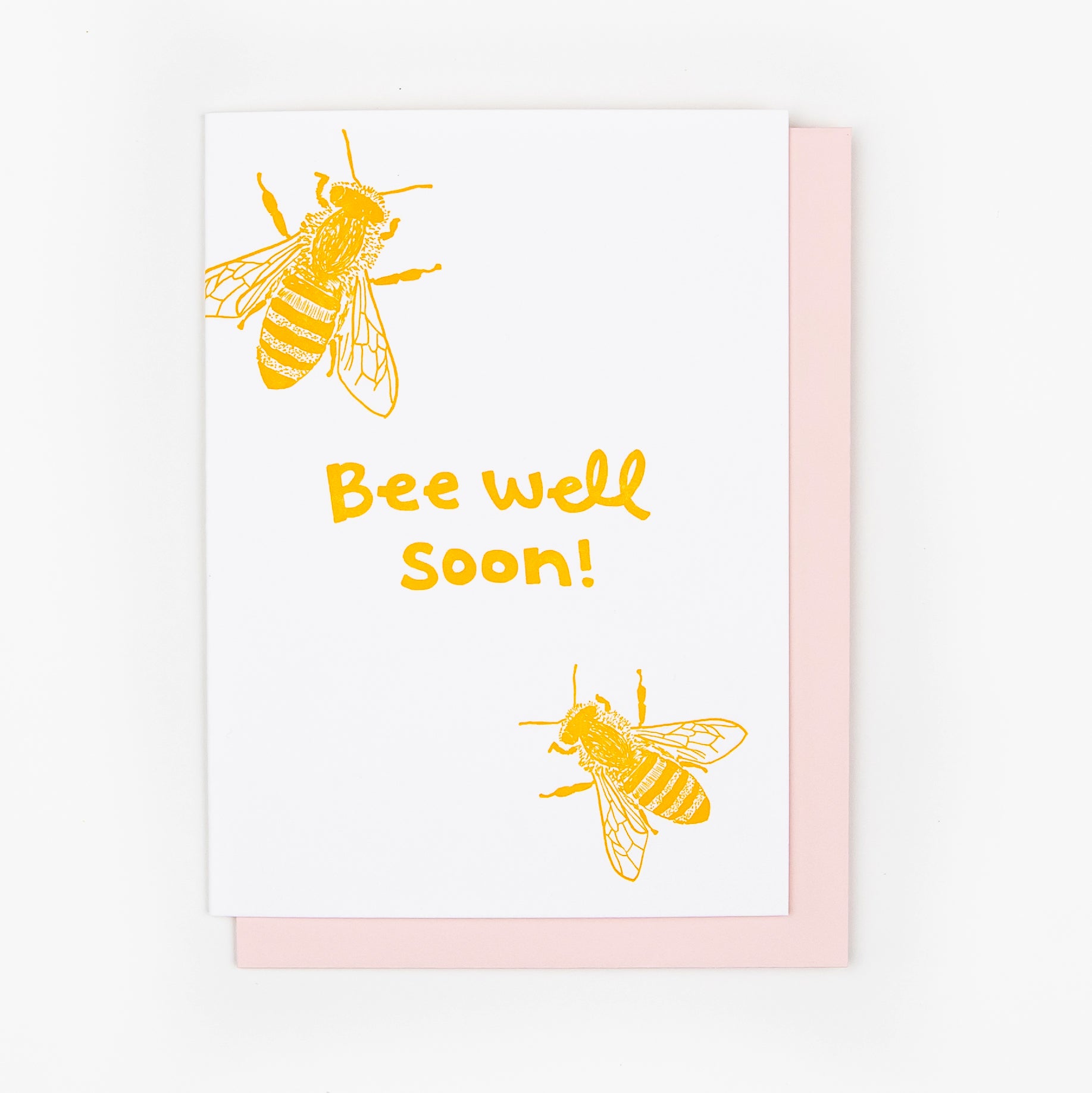 Letterpress greeting card featuring two hand-drawn honeybees, printed in a vibrant golden ink. Whimsical hand-drawn text saying "Bee Well Soon" is shown in the center of the card, in the same golden ink. The card is white, blank inside, and is paired with a pink envelope.