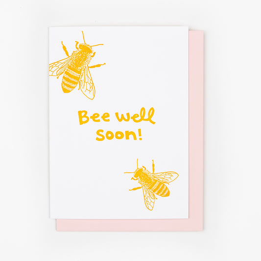 Letterpress greeting card featuring two hand-drawn honeybees, printed in a vibrant golden ink. Whimsical hand-drawn text saying "Bee Well Soon" is shown in the center of the card, in the same golden ink. The card is white, blank inside, and is paired with a pink envelope.