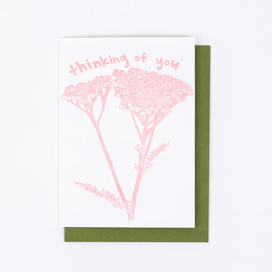 Letterpress greeting card featuring hand-drawn yarrow, printed in a rosy pink ink. Whimsical hand-drawn text saying "Thinking of You" is shown at the top of the card, in the same pink ink. The card is white, blank inside, and is paired with a green envelope.