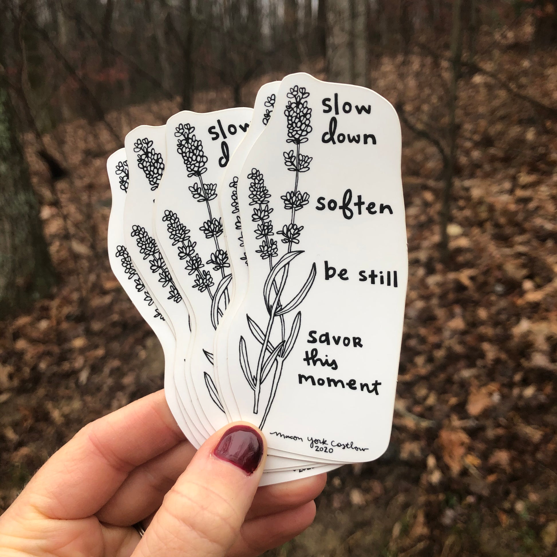 This Vinyl Sticker features original text by Macon York, hand drawn in her whimsical, folk art style. Hand-drawn lavender accompanies the text. The text reads "Slow Down. Soften. Be still. Savor this moment." A group of stickers fanned out is shown in front of a winter forest. 