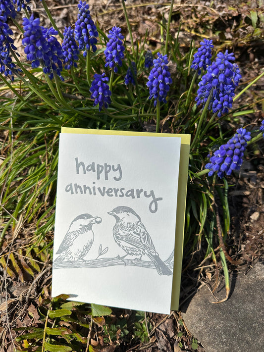 Letterpress greeting card featuring hand-drawn chickadees, printed in a shimmery silver ink. Whimsical hand-drawn text saying "Happy Anniversary" is shown on the top of the card, in the same silver ink. The card is white, blank inside, and is paired with a light yellow envelope. Card is shown outside on a sunny day in front of grape hyacinth flowers. 