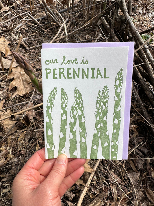 Letterpress greeting card featuring hand-drawn asparagus stalks, printed in a vibrant green ink. The top of the card says "Our love is perennial" in a whimsical hand-drawn text, in the same green ink. The card is white, blank inside, and is paired with a lilac purple envelope. Card is held up in an early spring garden bed of asparagus. 