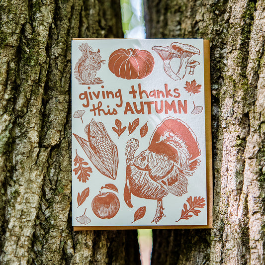 This cheerful Autumn card features all the iconic images of the fall season: squirrel, heritage pumpkin, mushrooms, fall leaves (maple, oak, sassafras, and ginkgo), a tom turkey, corn harvest, and a fresh-picked apple. Perfect for Thanksgiving. Vibrant orange ink paired with an orange envelope. Blank interior.  Card is shown outside by two trees. 