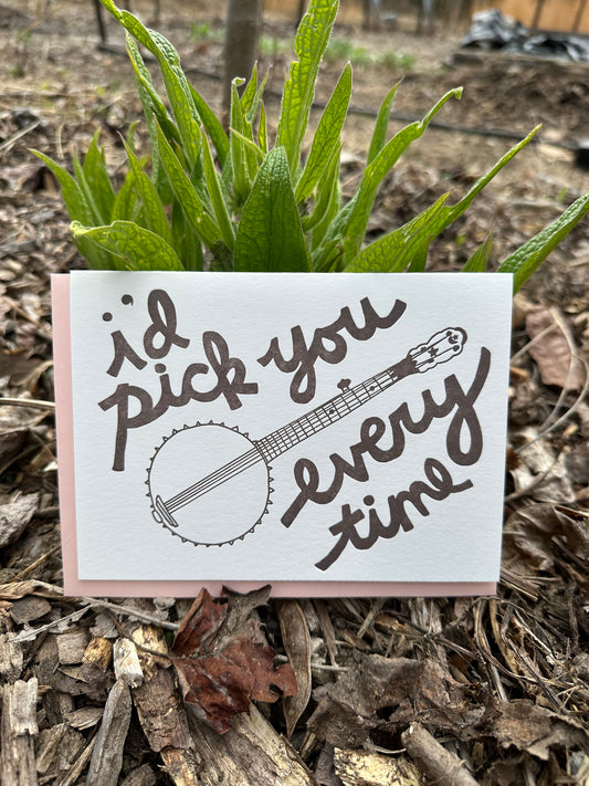 Letterpress greeting card featuring a hand-drawn banjo, printed in an earthy brown ink. Whimsical hand-drawn text saying "I'd Pick You Everytime" surrounds the banjo, in the same pink ink. The card is white, blank inside, and is paired with a light pink envelope. Card is shown in a garden bed in front of a comfrey plant. 